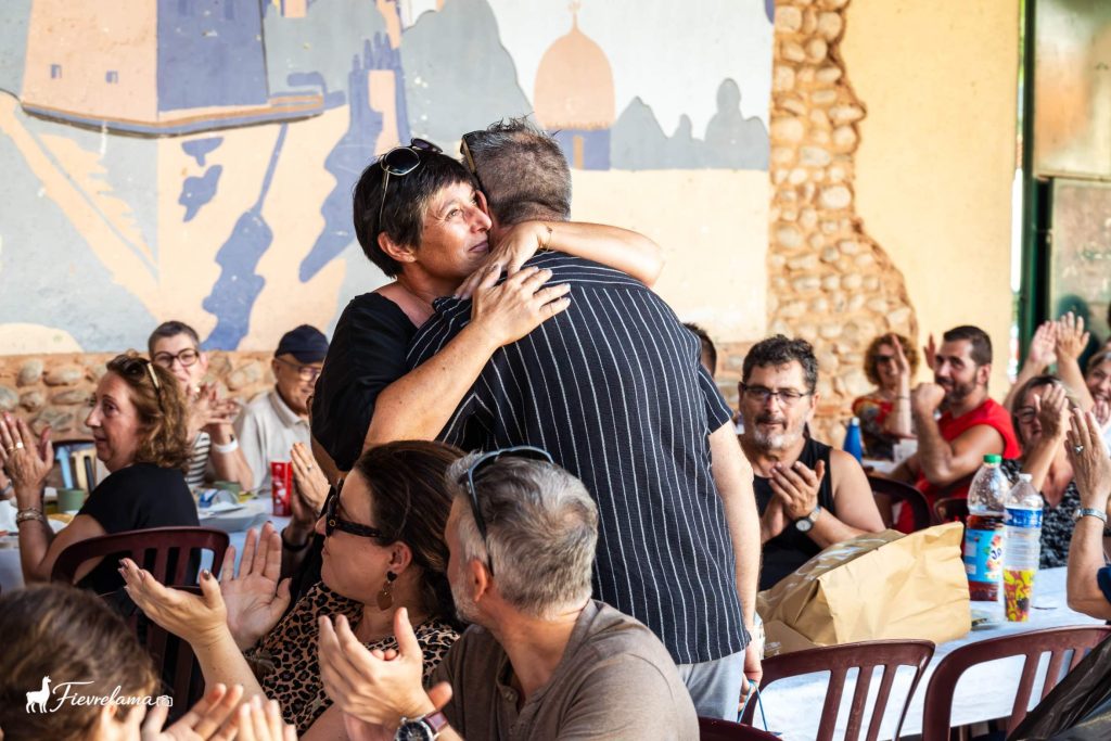Accolade émue entre deux membres sous les applaudissements des convives, devant la fresque du préau